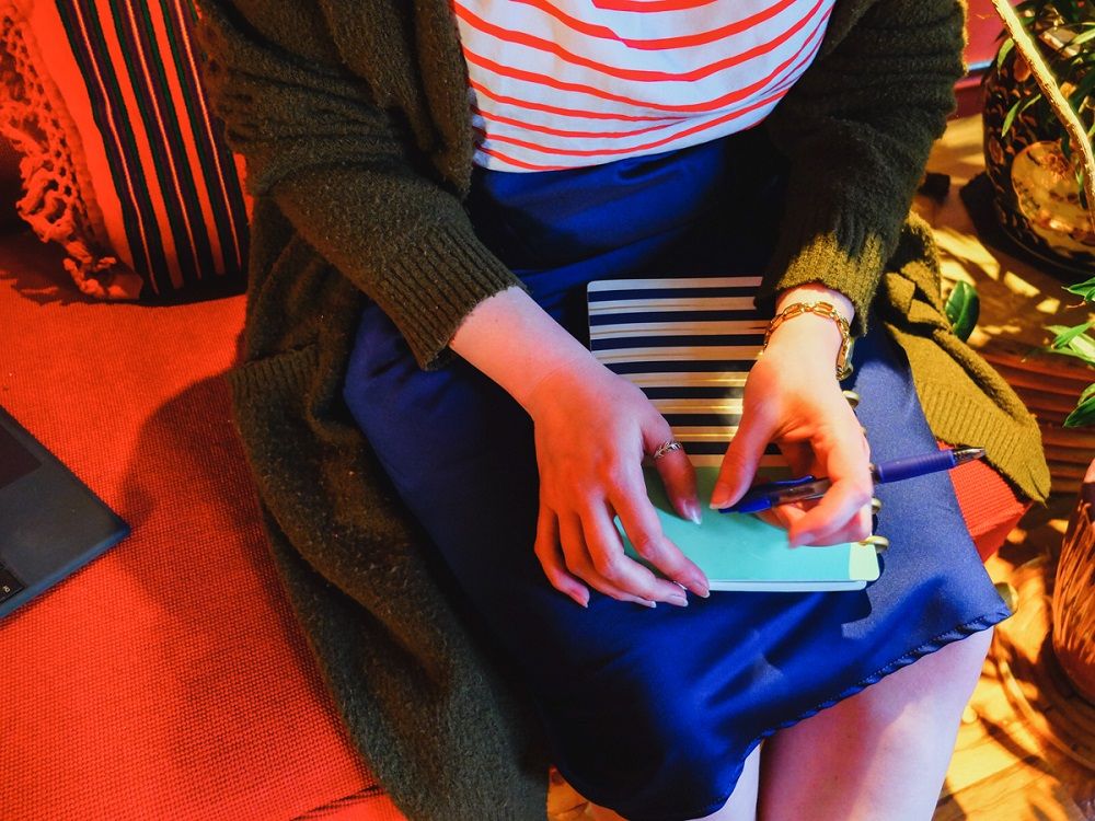 Leanna Lee sitting on a red chair in a striped shirt and blue skirt, holding a striped notebook and pen.