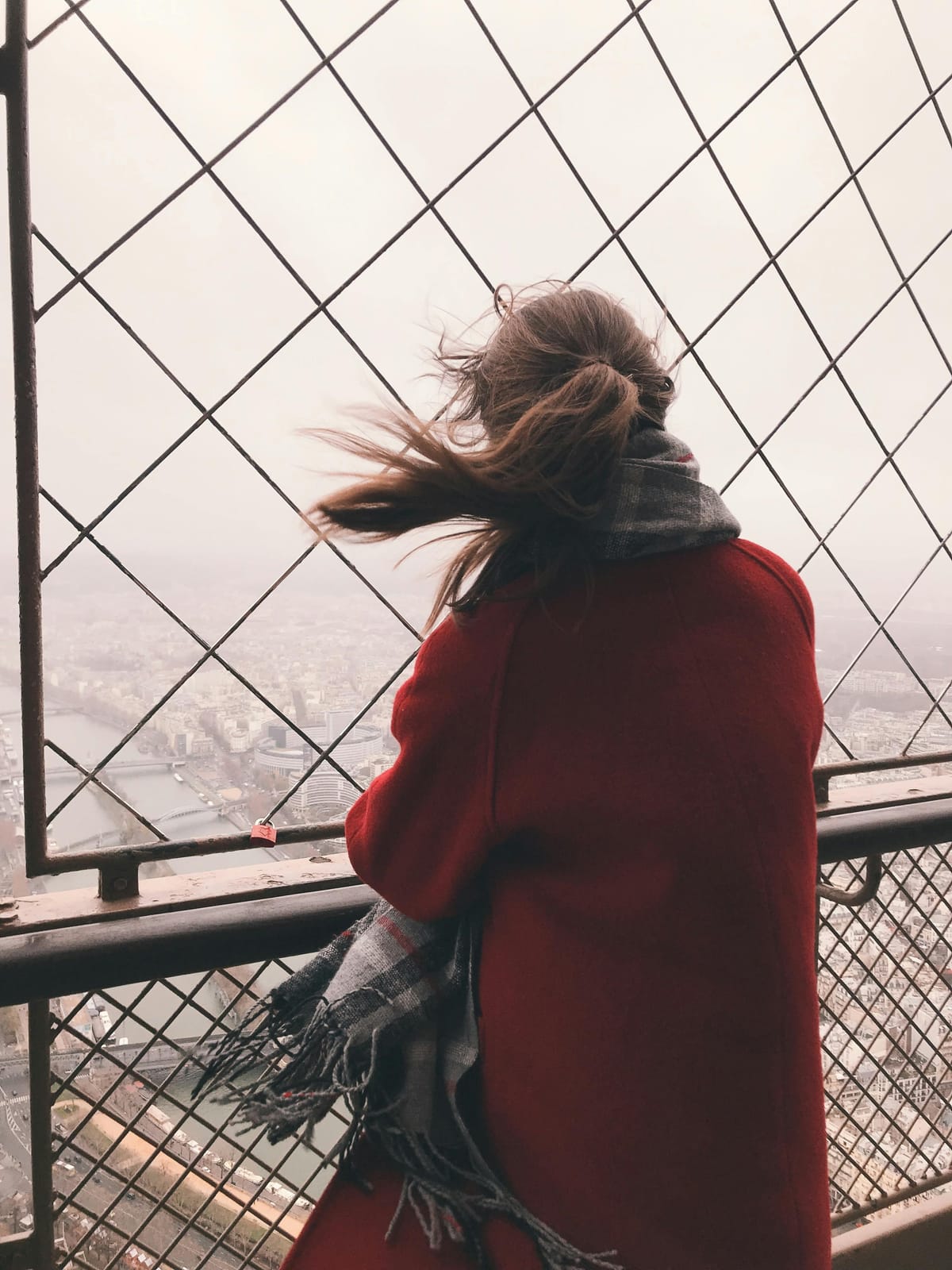 Woman in red coat and plaid scarf looking over a foggy city view through lattice bars as the wind whips her hair.