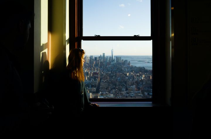Young woman with brown hair looks down at a cityscape out of upper story window awith the sunlight highlighting her face.