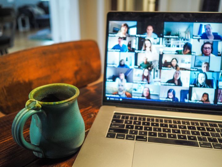 Open computer screen with lots of people in a video meeting on dark wooden table with a teal ceramic mug to one side.